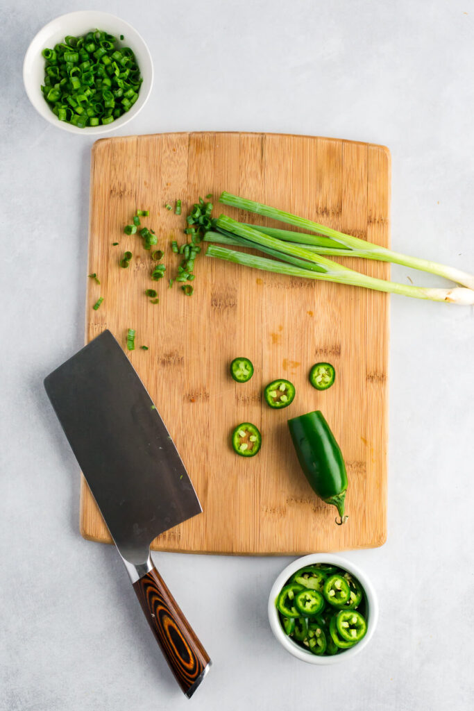 Jalapenos and green onions being chopped on a wooden cutting board 