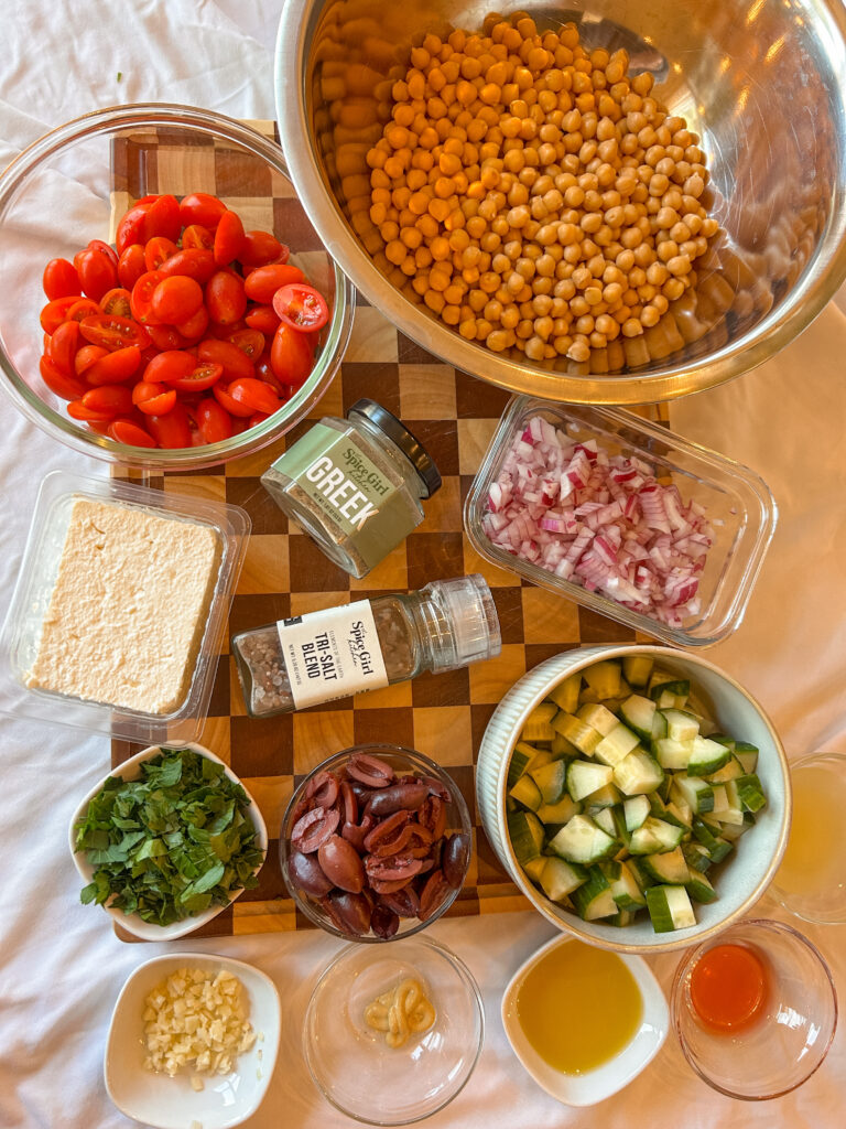 Ingredients needed to make Greek chickpea feta salad including chickpeas, feta, cucumber, tomatoes, olives, and Greek seasoning