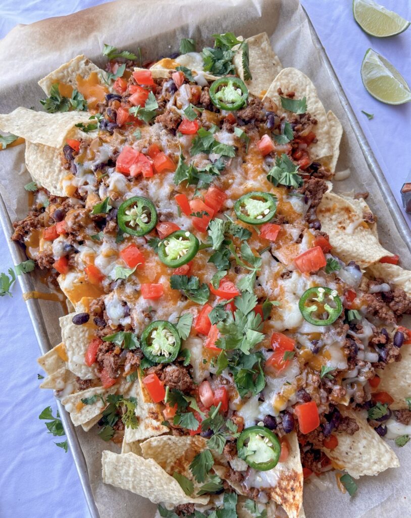 Sheet pan nachos finished with avocado, tomatoes, cilantro, jalapeños, sour cream, and lime wedges