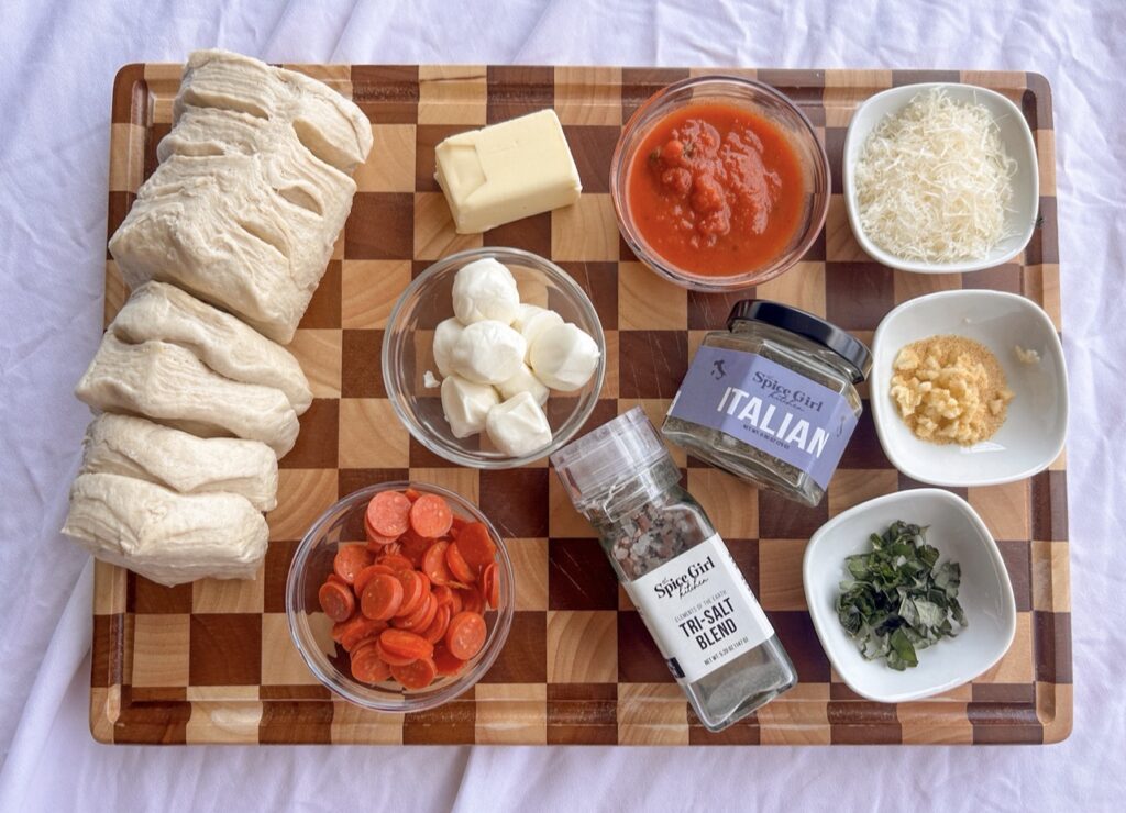 Ingredients for homemade pizza bombs including biscuit dough, pizza sauce, mini pepperoni, mozzarella, Parmesan, butter, garlic, parsley, and Italian seasoning.