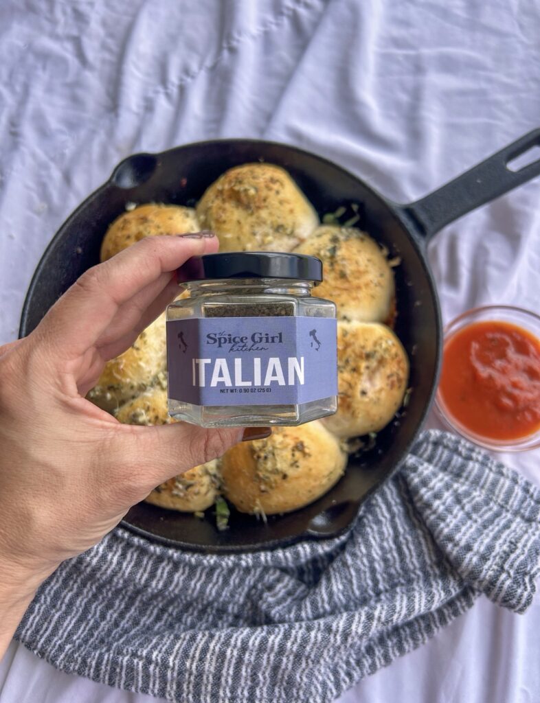 Homemade pizza bombs baked in a cast iron skillet with The Spice Girl Kitchen Italian Seasoning featured beside the finished recipe.