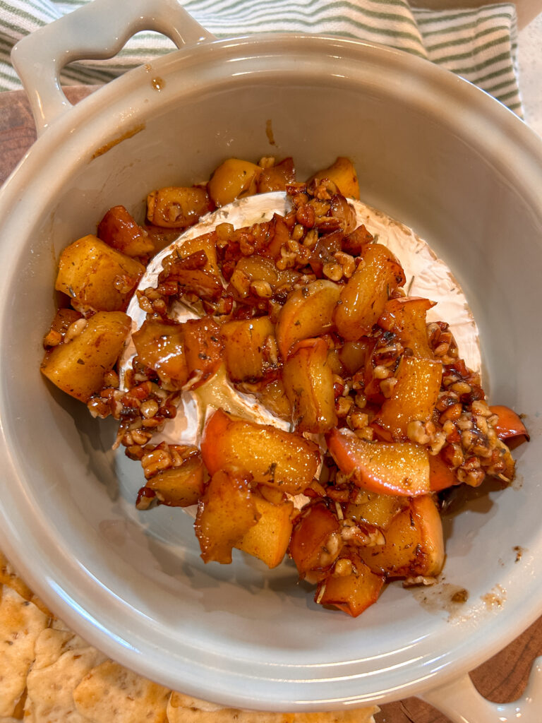 Cracker scooping warm baked Brie with caramelized apples and toasted pecans