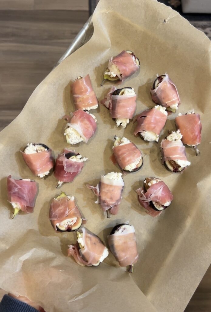 Halved fresh figs stuffed with Boursin cheese and wrapped in prosciutto on a baking sheet before baking.