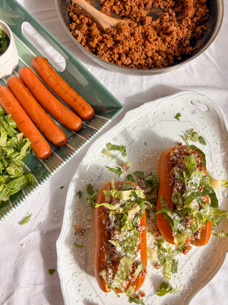 Assembling taco dogs with seasoned ground beef, shredded cheese, lettuce, tortilla chips, and salsa verde.
