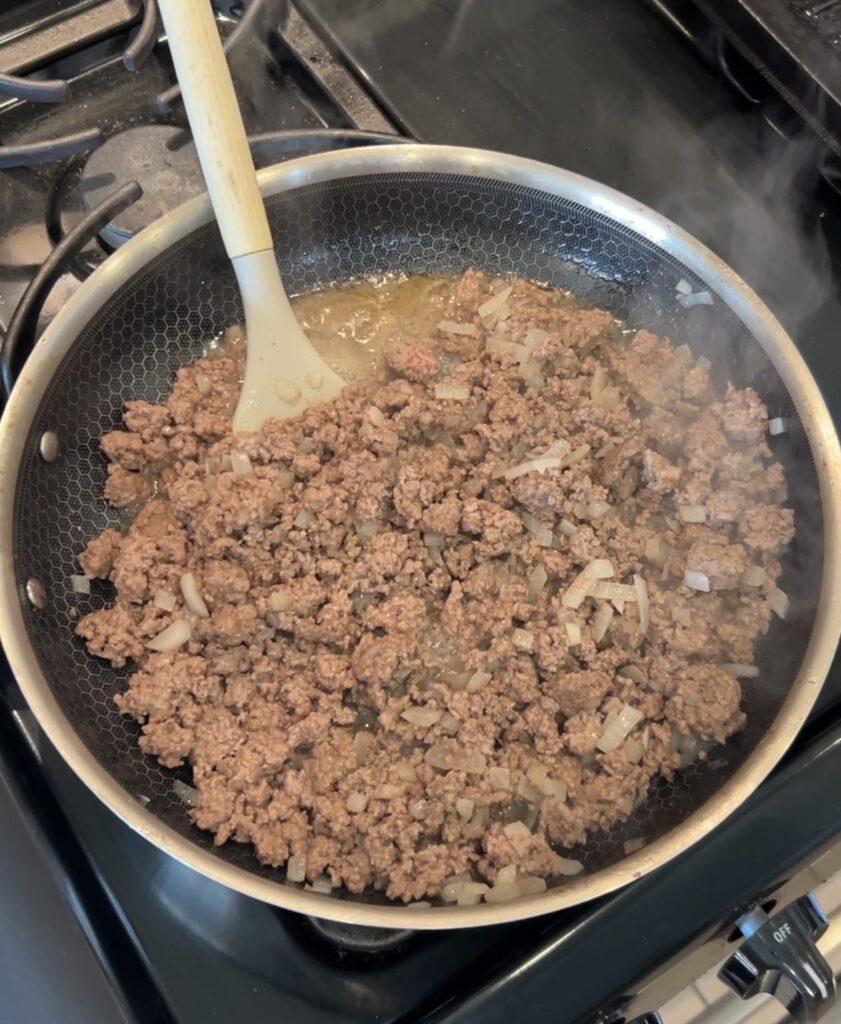 Ground beef and diced onion browning in a skillet to prepare the seasoned taco meat for taco dogs.