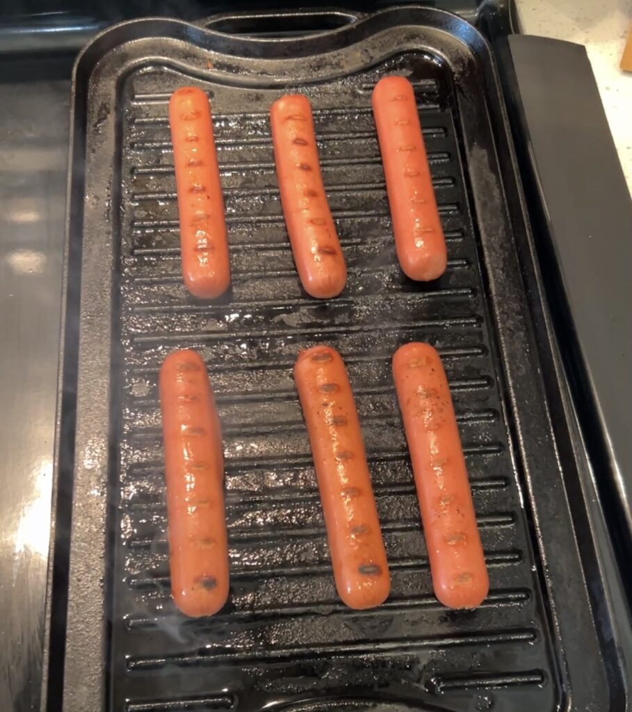 All-beef hot dogs cooking in a skillet until heated through and lightly browned for homemade taco dogs.