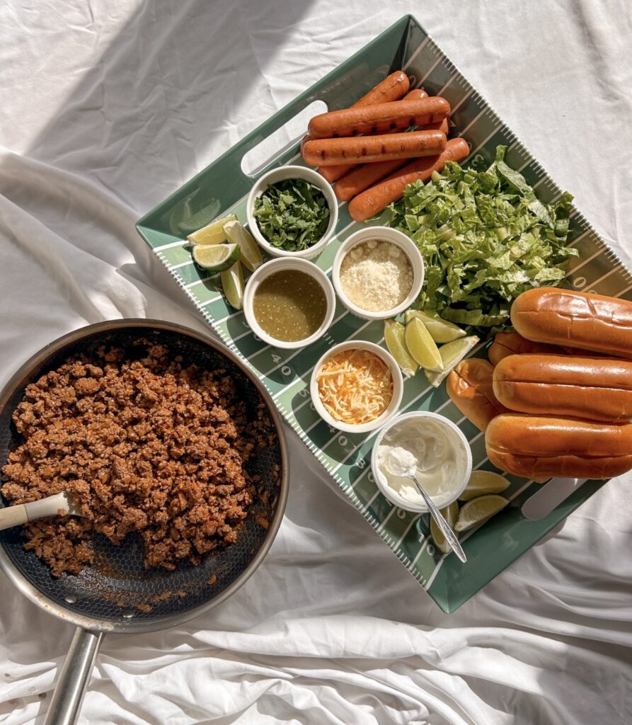 Ingredients for homemade taco dogs, including hot dogs, ground beef, Mexican seasoning, cheese, salsa verde, and fresh toppings.